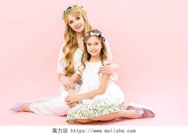 在粉色背景墙下的母女坐在地上美丽的女人和可爱的女儿在优雅的白色礼服和花卉花圈坐在地板上, 看着粉红色的背景相机